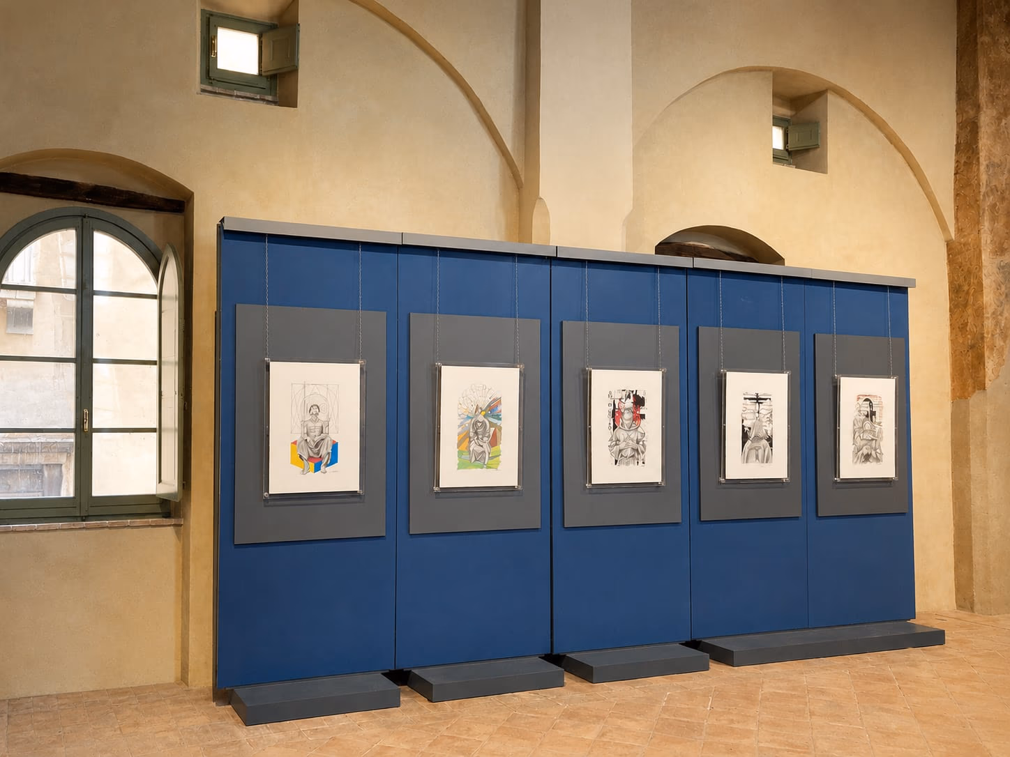 Una parete espositiva blu con cinque studi incorniciati, in una sala di Palazzo Trinci illuminata dalla finestra.