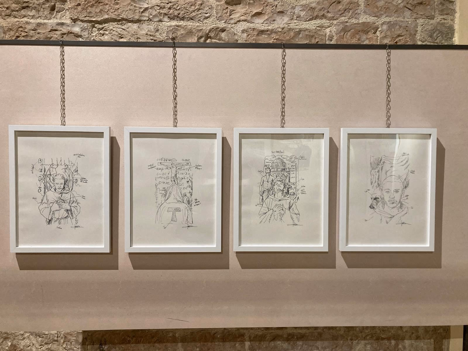 Four framed preparatory studies suspended on cables in front of the stone wall.