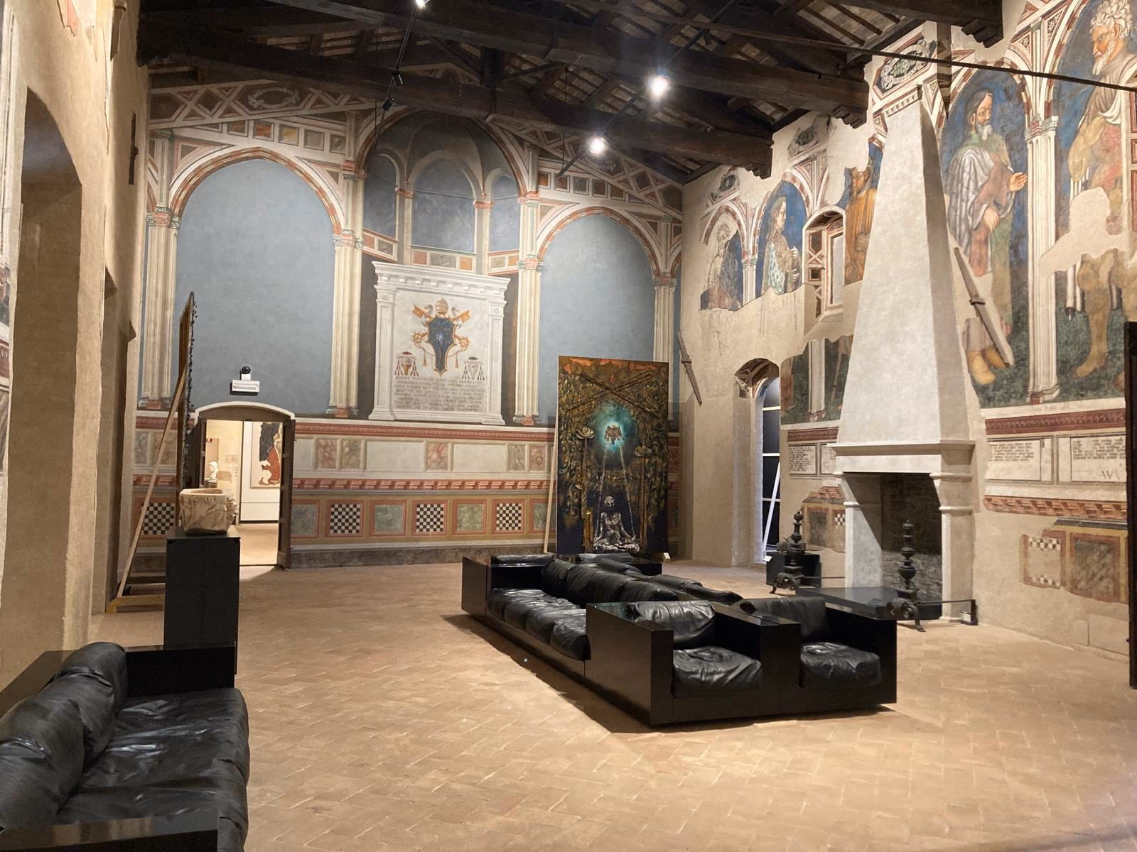 A large frescoed hall of Palazzo Trinci: frescoes on a blue ground, a tapestry on the wall and dark seating at the centre.