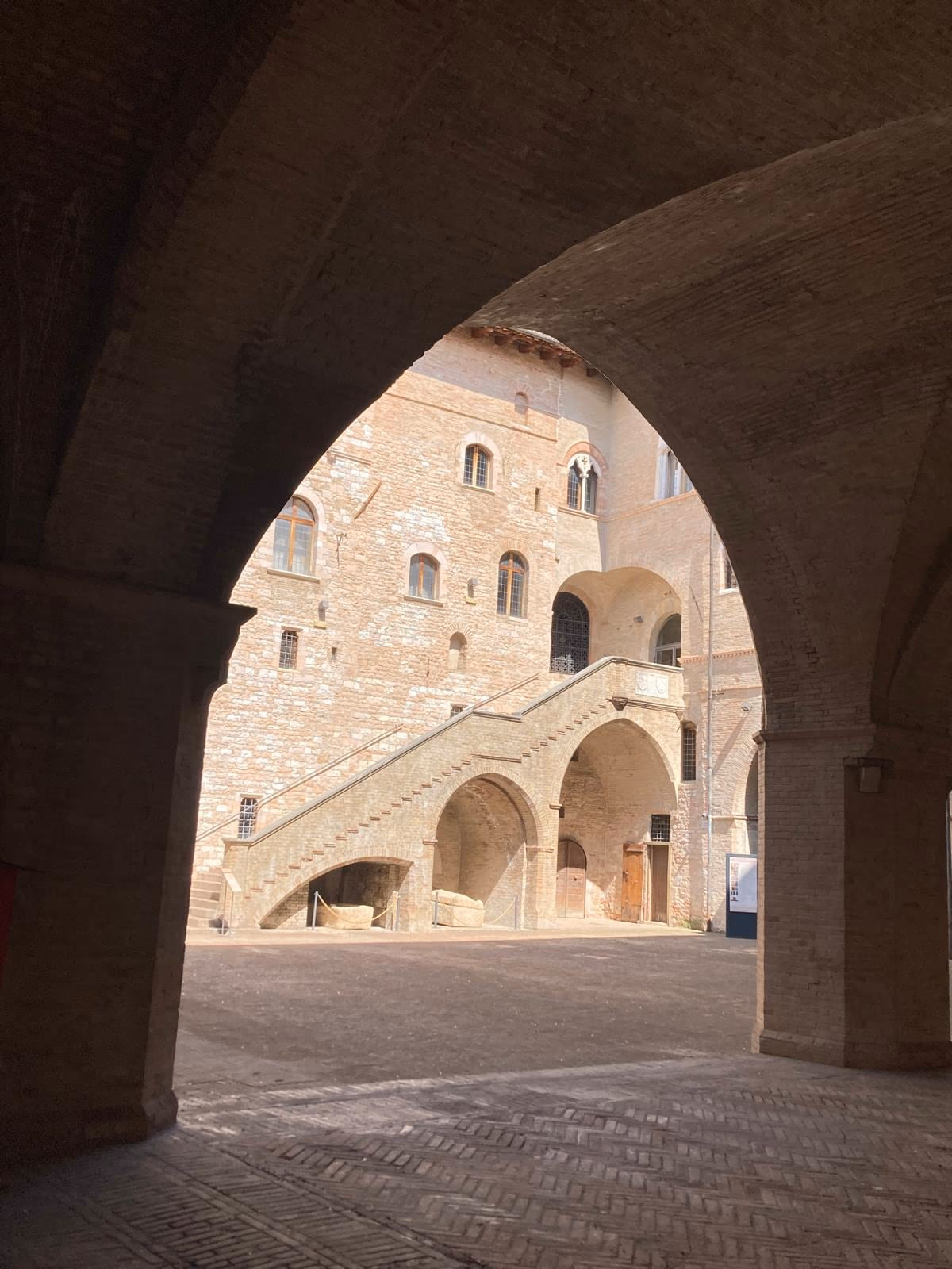 Il cortile di Palazzo Trinci visto attraverso un arco del porticato, con la scala esterna in pietra sul fondo.