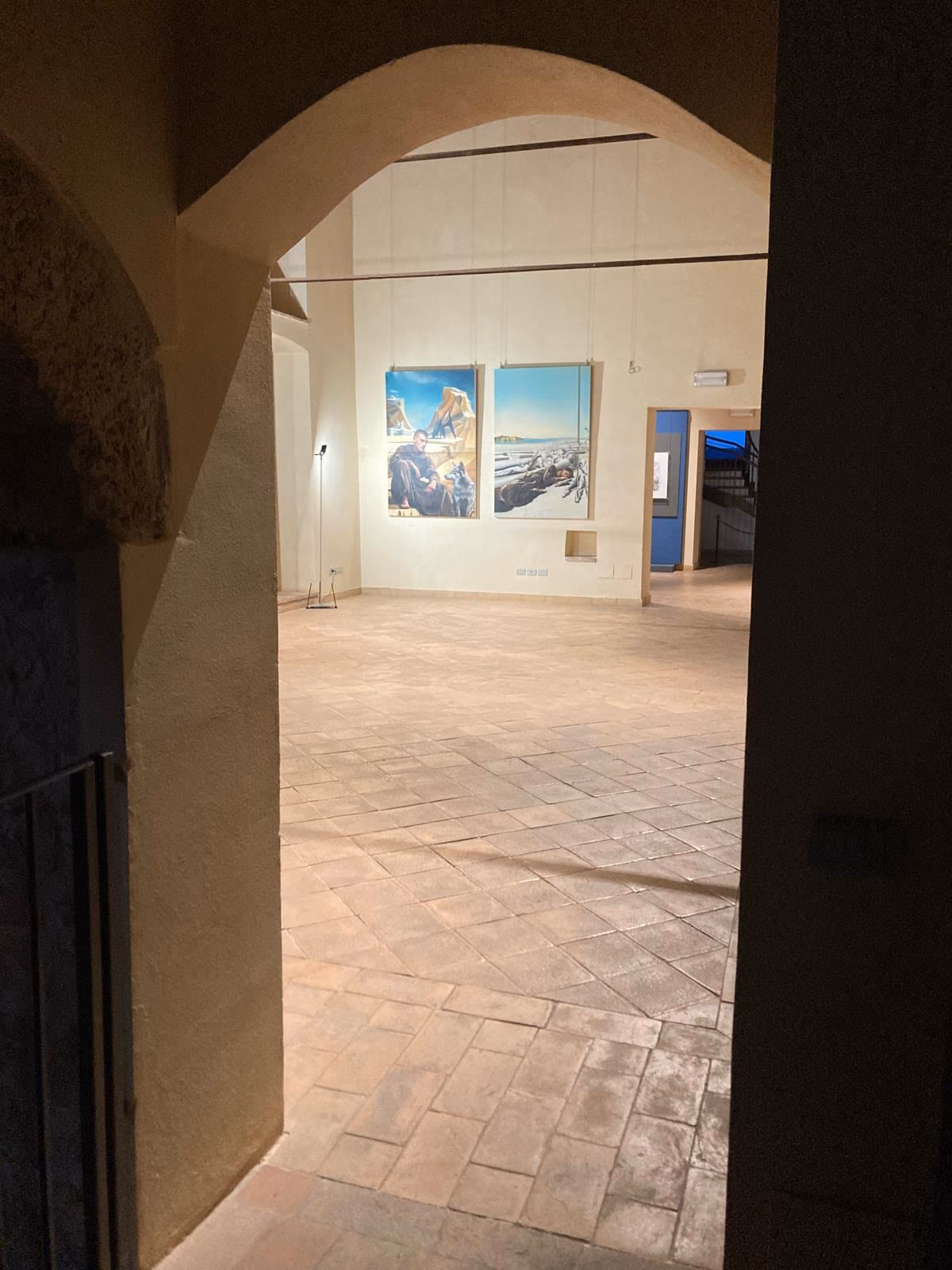 Un arco in pietra di Palazzo Trinci apre su una sala chiara dove sono appese opere incorniciate.